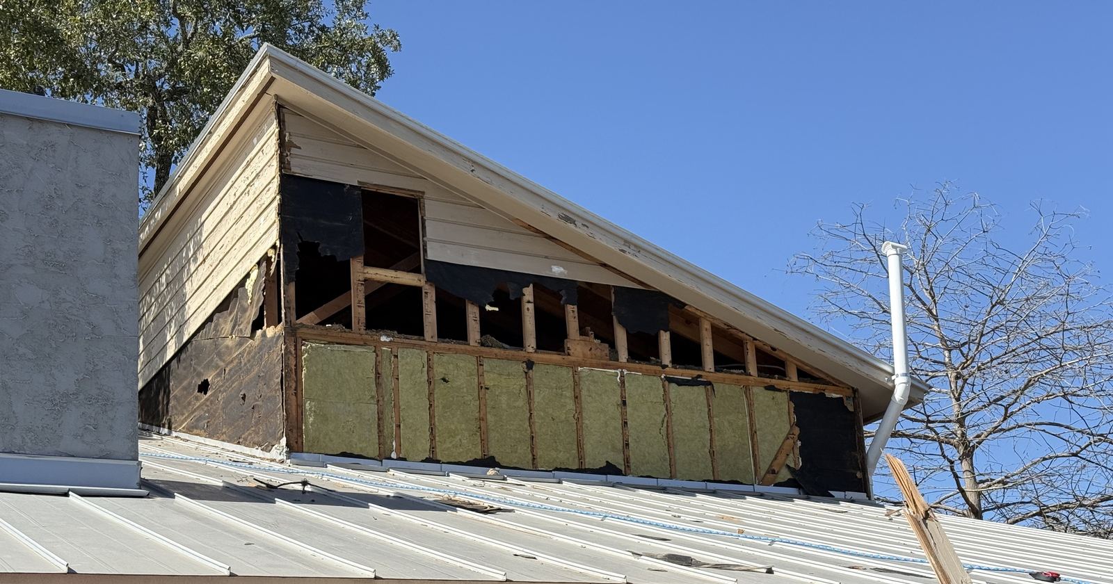 A gable wall with the siding torn open to the studs and the old metal roof below